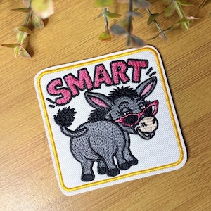 May include: A square embroidered patch with a white background and a yellow border. It features a gray donkey wearing pink glasses, looking back over its shoulder. Above the donkey, the word SMART is embroidered in bold, pink, capital letters.