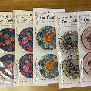 May include: A collection of car coasters with various designs, including sunflowers, floral patterns, and a highland cow. The coasters are round and packaged in clear plastic. Some coasters have text that reads "Be Free" and "Treat Yourself With Kindness".
