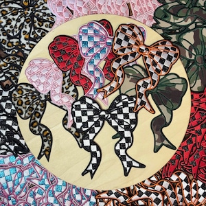 May include: A collection of decorative bow patches in various patterns and colors, including leopard print, checkered, and camouflage. The bows are arranged on a light wooden surface, showcasing their embroidered details and vibrant hues.