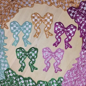 May include: A collection of decorative bow appliques in various colors, including gold, green, purple, and pink. Each bow features a checkered pattern with glitter accents. The bows are arranged on a light-colored surface, showcasing their intricate designs.