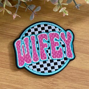 May include: A circular patch with the word "WIFEY" in pink, outlined in blue, and a black border. The background features a black and white checkered pattern. The patch is likely for clothing or accessories.