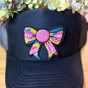 May include: Black trucker hat with a colorful embroidered bow patch. The bow features pink, yellow, and blue accents, with the word "DOUBLE" embroidered on the bow. The hat has a mesh back and a curved brim.