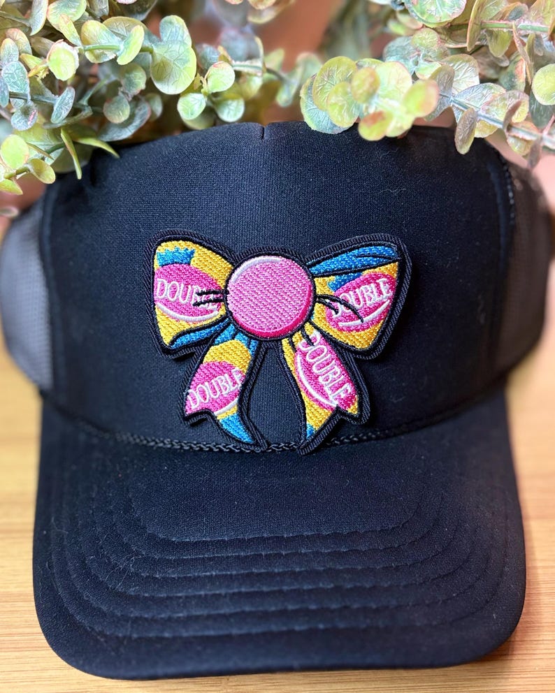 May include: Black trucker hat with a colorful embroidered bow patch. The bow features pink, yellow, and blue accents, with the word "DOUBLE" embroidered on the bow. The hat has a mesh back and a curved brim.
