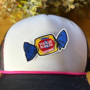 Pu&ograve; includere: Cappellino da camionista bianco con retro in rete nera e dettaglio corda rosa. Il cappello presenta una toppa di una confezione di caramelle gialla con la scritta "DOUBLES BUBBLES" in rosso e blu.