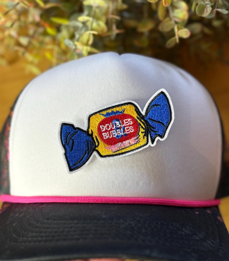 Pu&ograve; includere: Cappellino da camionista bianco con retro in rete nera e dettaglio corda rosa. Il cappello presenta una toppa di una confezione di caramelle gialla con la scritta "DOUBLES BUBBLES" in rosso e blu.