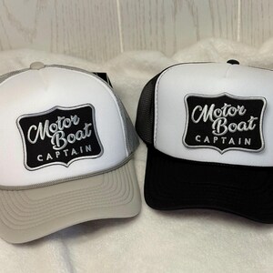 May include: Two trucker hats with white front panels and mesh backs. Each hat features a black patch with the words "Motor Boat Captain" in a stylized font. One hat has a gray brim and mesh, the other has a black brim and mesh.