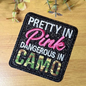 May include: A square embroidered patch with a black glitter background. The text reads PRETTY IN in white, Pink in cursive pink thread, DANGEROUS IN in white, and CAMO in a green, brown, and tan camouflage pattern with a black stitched border.