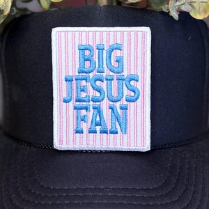May include: Black trucker hat with a mesh back and a patch that reads "BIG JESUS FAN" in blue lettering on a pink and white striped background. The hat has a curved brim and a black cord detail.