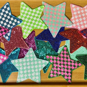 May include: A collection of star-shaped patches in various colors and patterns. The patches feature a checkered or dotted design in shades of pink, green, blue, red, and teal. The stars are arranged on a wooden surface.