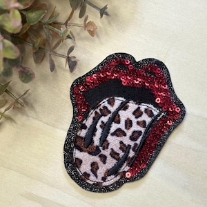 May include: A decorative patch in the shape of a mouth with a tongue sticking out. The mouth is outlined in black glitter and red sequins. The tongue has a leopard print design.