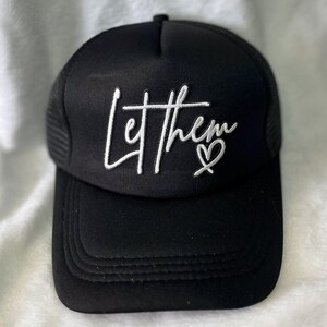 May include: Black trucker hat with a white embroidered design that says "Let them" with a heart symbol.