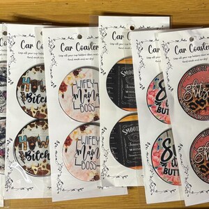May include: A collection of car coasters with various designs. Some feature floral patterns, skulls, and phrases like "Mama," "Wifey, Mama, Boss," and "How Bitch." Others display a bull skull or a whiskey label.