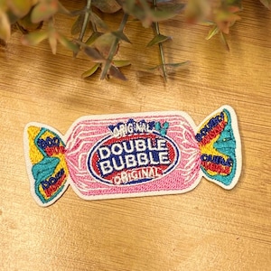 Pu&ograve; includere: Una toppa ricamata rosa e bianca a forma di caramella incartata. Presenta le parole "Double Bubble Original" in un ovale rosso e blu. Le estremit&agrave; della confezione sono gialle e turchese.