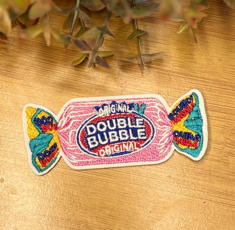 Pu&ograve; includere: Una toppa ricamata rosa e bianca a forma di caramella incartata. Presenta le parole "Double Bubble Original" in un ovale rosso e blu. Le estremit&agrave; della confezione sono gialle e turchese.