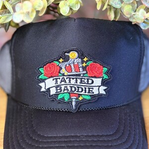 May include: Black trucker hat with a mesh back and a curved brim. The front features a patch with the words "TATTED BADDIE" in white, along with a tattoo machine, red roses, and gold accents. The hat is a stylish accessory.