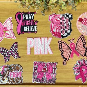 May include: A collection of pink embroidered patches with various designs, including butterflies, a bulldog, a boxing glove, and ribbons. Some patches feature text like "PRAY FIGHT BELIEVE" and "PINK OUT".