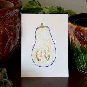 May include: A white greeting card featuring a colorful illustration of a sliced eggplant. The eggplant is outlined in blue, with green and yellow interior details. The card is displayed between two decorative ceramic pots.