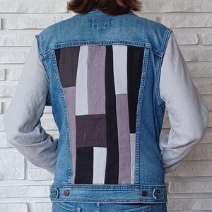 May include: A blue denim jacket with a patchwork design on the back. The patchwork is made of different shades of grey and black fabric. The jacket has long sleeves and a button closure.