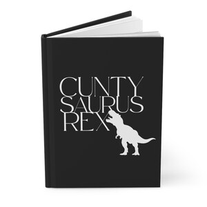 以下が含まれることがあります： 黒いハードカバーのジャーナル。白い文字で「CUNTY SAURUS REX」と、白いティラノサウルスレックスのシルエットが描かれています。ジャーナルは開いており、白いページが見えています。