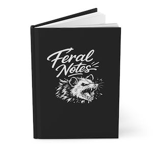 Peut inclure: Journal noir avec le texte "Feral Notes" en blanc au-dessus d'une illustration blanche d'un opossum grognant. Le journal est ouvert, révélant des pages lignées blanches.