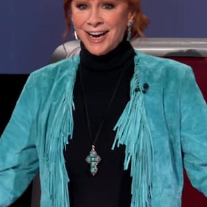 May include: A woman in a turquoise suede fringed jacket, black turtleneck, and cross pendant necklace. She wears a decorative belt buckle. Her auburn hair is styled. The background is a studio setting.
