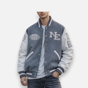 Puede incluir: Una chaqueta universitaria gris con mangas blancas, que presenta un logotipo NE blanco en el pecho, un emblema ovalado y el número 1960 en la manga. La chaqueta tiene cierre frontal con botones a presión, puños acanalados y cuello a rayas.