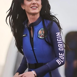 May include: A person wearing a blue and black jumpsuit with the words "BLUE ORIGIN" printed on the sleeve. The jumpsuit has a patch on the chest and a black belt. The person is smiling.