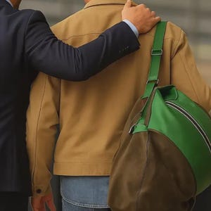 Puede incluir: Se ven una chaqueta color canela, vaqueros azules y una bolsa de lona verde y marr&oacute;n. Una persona con un traje azul marino tiene el brazo alrededor de la persona que lleva la chaqueta color canela. La bolsa de lona tiene una correa verde.