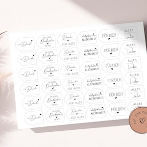 May include: A sheet of white stickers with black text and a heart design. The stickers have various phrases such as "Für Dich", "Ein kleines Dankeschön", and "Alles Liebe". A small, round sticker with text and a heart is in the bottom right corner.
