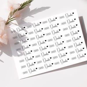 Puede incluir: Una hoja blanca de pegatinas con las palabras "Danke" y "Einfach" en escritura manuscrita, cada una acompañada de un pequeño corazón negro. Las pegatinas están dispuestas en un patrón repetitivo. Flores rosas en el fondo.