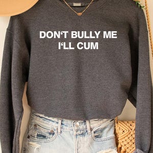 May include: A charcoal grey sweatshirt with the text "DON'T BULLY ME I'LL CUM" in white. The sweatshirt is paired with light blue denim shorts. A tan hat and a woven bag are also visible.