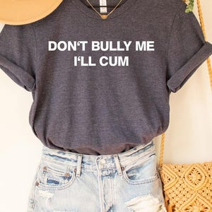 May include: A dark grey t-shirt with the text "DON'T BULLY ME I'LL CUM" in white. The shirt is paired with light blue denim shorts. A tan hat and a macrame bag are also visible.
