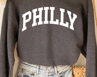 Puede incluir: Una sudadera gris oscuro con la palabra "PHILLY" en letras blancas. La sudadera se combina con pantalones cortos de mezclilla, un sombrero color canela y un bolso tejido. Los artículos se exhiben en una percha de madera contra una pared color crema.
