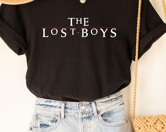 May include: Black t-shirt with white text that reads "THE LOST BOYS" with a red line under the text. The shirt is on a hanger with a straw hat and a straw bag.