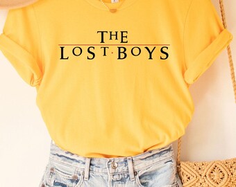May include: A yellow t-shirt with the text "THE LOST BOYS" printed on the front. The shirt is hanging on a wooden hanger and is styled with a straw hat and a woven bag.
