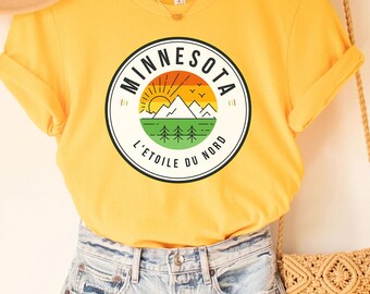 May include: A yellow t-shirt with a circular graphic featuring a mountain scene and the word "Minnesota". The shirt is paired with denim shorts, a tan hat, and a woven crossbody bag. The text "L'Etoile du Nord" is also visible.