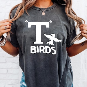 以下が含まれることがあります： 白い「T BIRDS」の文字と様式化された鳥のグラフィックが特徴の、濃い灰色のTシャツ。 ヴィンテージ感のあるウォッシュ加工で、半袖です。 カジュアルなユニセックスTシャツ。