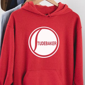 Könnte beinhalten: Roter Kapuzenpullover mit einem weißen Kreislogo, auf dem "Studebaker" in weißer Schrift steht.
