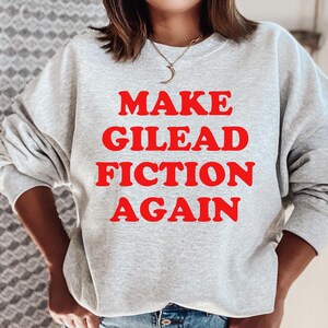 Può includere: Felpa grigia con la scritta "Make Gilead Fiction Again" stampata in rosso.