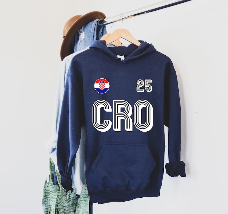 May include: Navy blue hoodie with a Croatian flag emblem and the number 25. The word "CRO" is printed in large white letters. The hoodie has a front pocket and a drawstring hood.