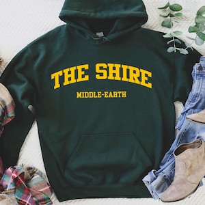 May include: A dark green hooded sweatshirt with the text "THE SHIRE" in yellow arched lettering and "MIDDLE-EARTH" in smaller yellow lettering below.