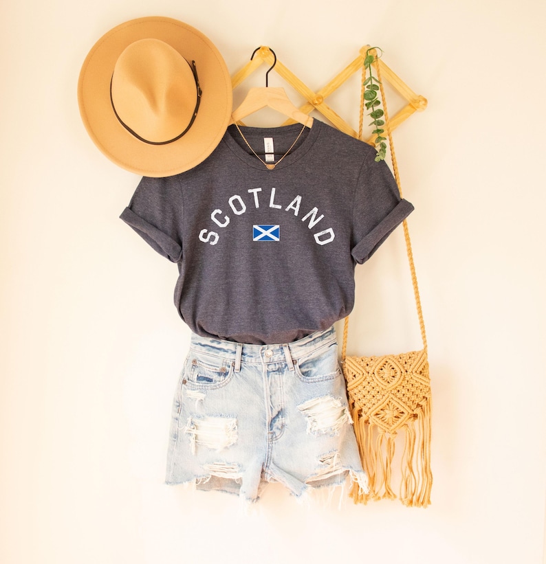 May include: A dark gray t-shirt with the word "SCOTLAND" in white arched lettering and a small Scottish flag graphic. The shirt is paired with light blue denim shorts, a tan hat, and a woven crossbody bag.