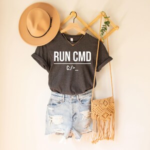 Op de afbeelding: Een donkergrijs t-shirt met de tekst "RUN CMD" en "C:/>_" in het wit. Het shirt wordt gecombineerd met lichtblauwe spijkerbroekshorts, een bruine hoed en een geweven crossbodytas met franjes.