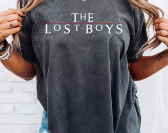 Può includere: T-shirt grigio scuro con la scritta bianca "THE LOST BOYS", con una linea rossa sotto "THE". La maglietta a maniche corte ha un girocollo. Il tessuto sembra morbido e confortevole.