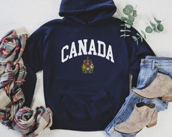 Puede incluir: Sudadera con capucha azul marino con la palabra "CANADA" en letras blancas arqueadas. Debajo, un emblema colorido. La imagen también muestra vaqueros azules, botas color canela y una bufanda a cuadros. Un atuendo casual.