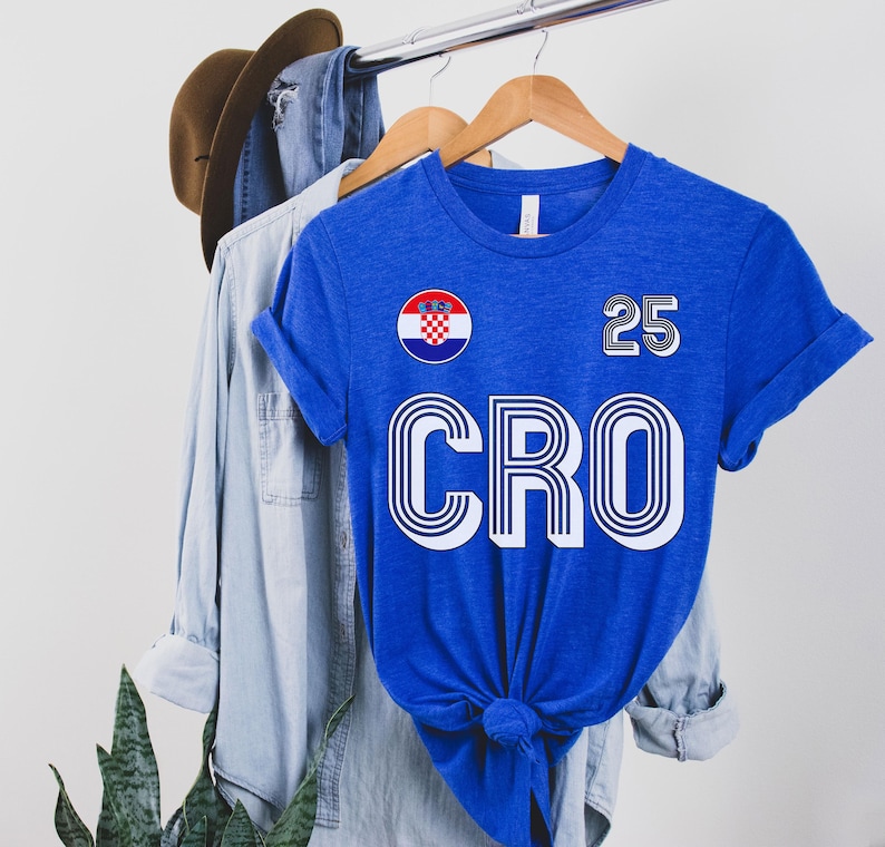 Pu&ograve; includere: Una maglietta blu reale con una grafica "CRO" bianca e nera e il numero "25" sul davanti. La maglietta presenta anche una toppa circolare con la bandiera croata.