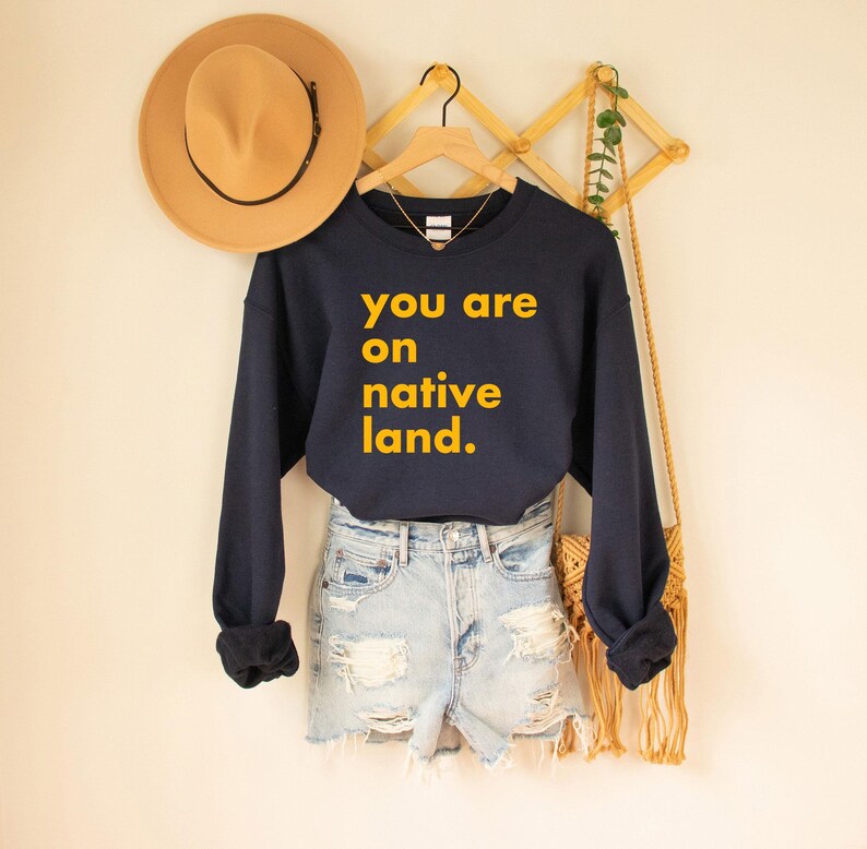 Op de afbeelding: Een marineblauw sweatshirt met de tekst "you are on native land" in het geel. Het sweatshirt wordt gecombineerd met een lichtblauwe spijkerbroek. Een bruine hoed en een geweven tas hangen aan een houten rek.