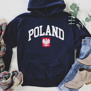 May include: Navy blue hooded sweatshirt with the word "POLAND" in white arched lettering. Below the text is a red and white Polish coat of arms. The sweatshirt is paired with blue jeans, tan boots, and a plaid scarf.