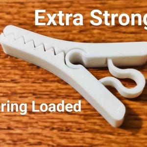 Może przedstawiać: Biały plastikowy zacisk sprężynowy z etykietą "Extra Strong" i "Spring Loaded".