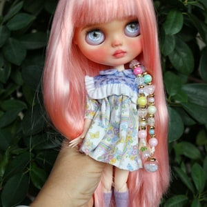 May include: A custom Blythe doll with long pink hair and large blue eyes. The doll wears a floral print dress, lavender socks, and blue shoes. A beaded accessory hangs from the doll's hair. The doll is held in front of a green leafy background.